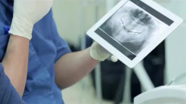 Modern bir diş kliniğinde, önlüklü bir diş hekimi bir tablette hastanın dişlerinin röntgenini dikkatle gösterir. Bu yakın çekim diş bakımının karmaşık detaylarını yakalar ve teknolojinin sözlü sağlık değerlendirmelerindeki önemini vurgular.