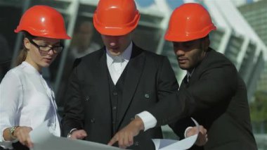 Üç kendine güvenen mimar, grevdeki turuncu kasklarla süslenmiş, açık havada duruyor, dikkatle planlarını gözden geçiriyorlar. Odaklanmış ifadeleri ve işbirlikçi ruhları, inşaat dünyasının dinamik dünyasında takım çalışmasının özünü vurgular.