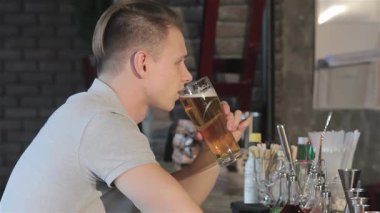 Neşeli sarışın bir adam barda oturuyor, birasından bir yudum aldıktan sonra sırıtıyor. Sıcak atmosfer, kırsal tuğla bir duvar ve ışıldayan bir bar takımı ile rahatlama ve dostluk manzarasını geliştirir.