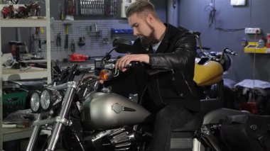 Tarz sahibi genç bir erkek motorcu, hareketli bir atölyede aletler ve motorsiklet parçalarıyla çevrili klasik helikopterinde güven içinde poz veriyor. Çekici beyaz adam, bakımlı sakallı ve parlak siyah deri ceketli, ustalıkla dokunuyor.