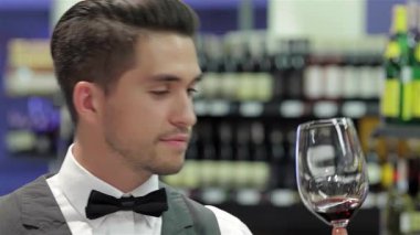 Şık bir smokin giymiş dengeli bir erkek şarap garsonu, kırsal bir ahşap masaya yaslanmış bir şarap kadehini keskin bir gözle inceliyor. Arka planda zarif bir şarap rafı yer alıyor, sofistike olanları geliştiren zengin bir şişe koleksiyonu sergileniyor.