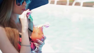 Geniş kenarlı koyu mavi şapkalı çarpıcı bir kadın ferahlatıcı bir kokteylin tadını çıkarıyor, neşeli gülüşü neşe saçıyor. Güneş gözlükleriyle güneşi yansıtan bu çekici kahverengi saçlı kadın yaz tatili ve rahatlığının özünü temsil ediyor.