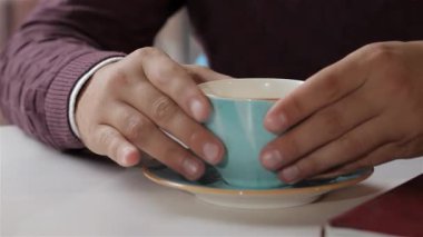 Yakın plan çekimde bir çift erkek eli zarif bir şekilde mavi kahve fincanını narin tabağından kaldırıyor. Sahne sıcaklığı ve rahatlığı yansıtıyor, kahveyi sevenleri zengin aromanın ve en sevdikleri biranın tadını almaya davet ediyor.