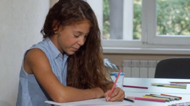 Neşeli, uzun saçlı bir kız okulda çizimine dalmış, kameraya gülümserken mutluluk saçıyor. Bu hoş sahne onun yaratıcılığını ve coşkusunu yakalıyor. Renkli sanat malzemeleriyle dolu canlı bir atmosfer sergiliyor.