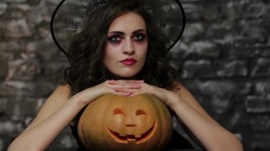 Büyücü bir kadını somutlaştıran büyüleyici kadın büyüleyici bir poz veriyor sırıtan bir balkabağıyla siyah, desenli tuğla bir duvara vuruyor. Dramatik makyajı ve gotik kıyafetleri ürkütücü Cadılar Bayramı atmosferini güçlendiriyor ve izleyicileri sihir dünyasına davet ediyor.