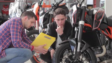 Canlı bir motosiklet galerisinde, siyah ceketli genç bir adam yoğun bir şekilde kareli gömlekli bir danışman gibi dinleyerek çarpıcı siyah spor bir motosikletin özelliklerini açıklar. Motosikletleri tartışırken ortam heyecanla uğulduyor.