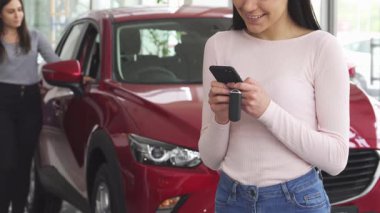 Neşeli bir kadın akıllı telefonuna mesajlar yazarken gülümsüyor, galeride çarpıcı kırmızı bir arabanın önünde heyecanını gösteriyor. Arka planda, başka bir bayan müşteri otomobili inceler ve arabanın canlı atmosferini ekler.
