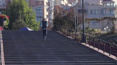 Kendini adamış bir sporcu, renkli sonbahar yapraklarıyla çevrili manzaralı bir şehir parkının merdivenlerinde enerjik bir şekilde koşuyor. Bu canlı sahne zindelik ve canlılığın özünü yakalar, modern bir arka planda açık hava egzersizinin neşesini gözler önüne serer