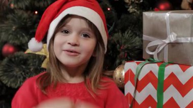 Noel Baba şapkası takan sevimli küçük bir kız Noel için heyecanını ifade ederken saf neşe saçıyor. Güzelce paketlenmiş hediyeler ve ışıl ışıl bir Noel ağacı ile çevrili neşeli çığlıkları ve kaldırdığı eller,