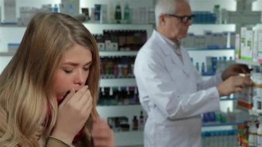 Uzun sarı saçlı ve çarpıcı kırmızı atkısı olan güzel beyaz bir kadın yavaşça öksürüyor. Eczanede ilaç ararken endişeli görünüyor. Arka planda, beyaz önlüklü odaklanmış bir erkek eczacı özenle ürünleri düzenliyor.