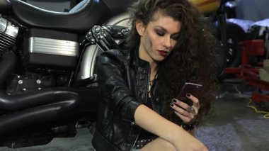 Kıvırcık saçlı çekici bir beyaz kadın atölye zemininde oturmuş, akıllı telefonuyla çarpıcı bir motorsikletin yanında neşeyle sohbet ediyor. Şık, siyah bir deri ceket ve kot şort giymişti. Kendine güveni ve tutkusu vardı.