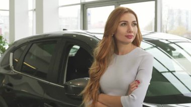 Çarpıcı kızıl saçlı bir kadın, modern bir bayilikte zarif yeni arabasına yaslanırken mutluluk yayıyor. Düşünceli bakışları ve zarif duruşu sahipliğin heyecanını, başarıyı ve yeni maceraların heyecanını yansıtıyor.