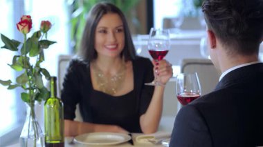 Büyüleyici genç bir çift romantik bir akşam yemeğinden zevk alır, kırmızı şarap kadehleriyle kadeh kaldırırken sevgiyle gülümserler. Güllerle süslenmiş şık restoran ortamı, özel bir olayı kutlamak için mükemmel bir ortam sağlıyor.