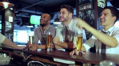 Üç hevesli arkadaş, şık günlük kıyafetler giymiş, neşeli bir şekilde tezahürat yapıyor ve canlı bir barda yoğun bir futbol maçı izlerken bardaklarını kaldırıyorlar. Atmosfer heyecan verici, kahkaha ve dostluk dolu.