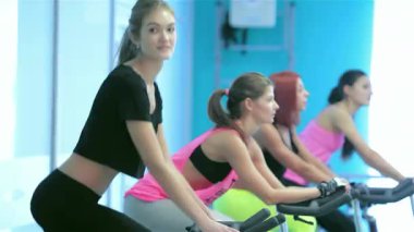 Hareketli bir spor salonu ortamında, atletik bir kız sabit bir bisiklete güvenle pedal çeviriyor, kameraya parlak bir gülümseme gösteriyor. Odaklanmış enerjisi arka planda güçlü bir şekilde pedal çeviren kız arkadaşlarıyla çelişiyor.