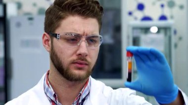 Gıcır gıcır beyaz laboratuvar önlüklü yakışıklı esmer bir adam renkli bir test tüpünü inceliyor ve biyoteknoloji uzmanlığını sergiliyor. Tüpün içindeki canlı sıvı onun konsantrasyonunu ve kararlılığını yansıtıyor.