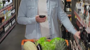 Şık sakallı çekici bir beyaz adam, kalabalık bir süpermarket koridorunda şişeyle bira kutusu arasında ince eleyip sık dokuyor. Etrafı çeşitli içeceklerle dolu renkli raflarla çevrili bir elinde akıllı bir telefonu dengeliyor.