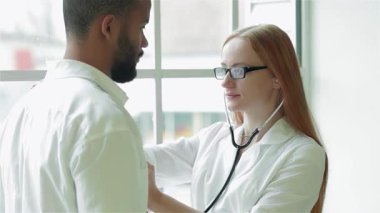 Parlak ve modern bir klinikte, şefkatli bir bayan doktor steteskopuyla genç bir adamın göğsünü dikkatle dinler ve kapsamlı bir muayene sağlar. Sıcak atmosfer profesyonel etkileşimlerini vurguluyor.