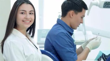 Parlak ve neşeli bir dişçi yardımcısı kendini işine adamış bir dişçinin yanına oturmuş sıcak bir gülümseme yayıyor. Bu canlı sahne modern bir diş kliniğinde takım çalışmasının özünü yakalıyor, profesyonellik ve dostluk gösterisi sergiliyor.