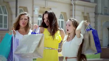 Üç modaya uygun kız ellerinde renkli alışveriş torbaları, sokakta yürürken mutluluk saçıyorlar, kahkahaları havada yankılanıyor. Parlak atmosfer, kaygısız ifadeleriyle ve aydınlatıcı güneş ışığıyla güçlenir.