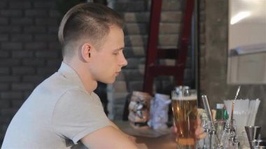 Yakışıklı, genç, beyaz bir adam modern bir barda oturuyor ve uzun bir bardak altın birayı dudaklarına kaldırıyor. Kırsal tuğla bir duvarla vurgulanan ılık ortam, uzun bir günün ardından gevşemek için mükemmel bir atmosfer yaratıyor.