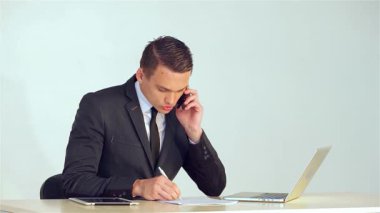 Sivri takım elbiseli kararlı genç bir işadamı, bir telefon görüşmesinde notlar alırken dikkatle not alıyor. Şık dizüstü bilgisayarı ve tableti modern masanın üzerinde duruyor. Profesyonelliği somutlaştıran dinamik bir çalışma alanı yaratıyor.