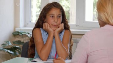 Uzun, dalgalı saçları olan heyecanlı bir genç kız, öğretmeniyle hevesle konuşurken çenesini elleriyle dinlendirirken öne doğru eğiliyor. Doğal ışıkla dolu parlak sınıf ortamı, sıcak bir öğrenme ve bağlantı atmosferi yayıyor.