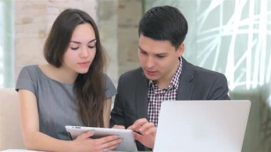 Genç, çekici bir iş adamı ve kadın, bir tablet üzerindeki verileri incelerken, kameraya içten bir şekilde gülümseyen kadınla canlı bir tartışmaya girişirler. Profesyonel kıyafetleri ve odaklanmış ifadeleri takım çalışması ve yenilik ortamı yaratıyor.