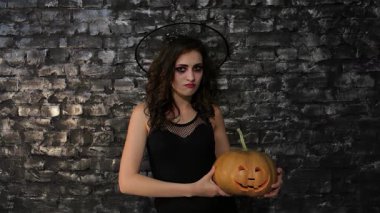 Çekici bir kadın büyücü, parlak siyah bir kıyafet giymiş, yaramaz bir sırıtışla oyulmuş bir balkabağı tutuyor, desenli siyah tuğla bir duvara dayanıyor. Bu Cadılar Bayramı 'ndan esinlenilmiş görüntü bir gizem ve sihir havası yayıyor.