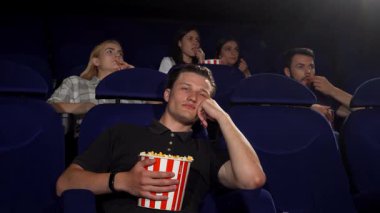 Rahat bir ifadeyle genç, çekici bir adam lüks bir sinema koltuğunda elinde patlamış mısırla uyukluyor. Onu çevreleyen büyüleyici bir seyirci ekranı seyrediyor. Uykulu tavrı ve onların nişanlı tavırları arasında katı bir kontrast oluşturuyor.