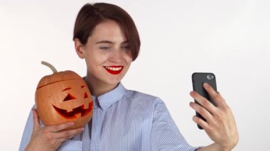 Kırmızı dudaklı çekici genç bir kadın neşeli bir şekilde Cadılar Bayramı balkabağıyla selfie çekerek neşeli ruhunu sergiliyor. Yakın tutulan canlı kriko-fener, bu keyifli ana neşeli bir dokunuş ekler, karıştırıcı teknoloji ve