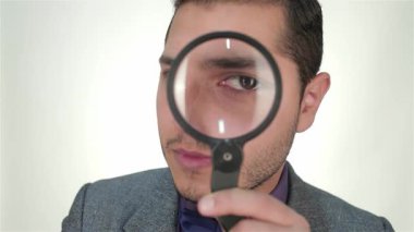 Odaklanmış genç bir işadamı büyüteçten bakıp karmaşık detaylarla dolu bir dünyayı gözler önüne seriyor. Kendine güveni ve keskin takım elbisesi, profesyonel havayı geliştiren temiz beyaz bir arkaplan karşısında kararlılık ve başarı gösteriyor.