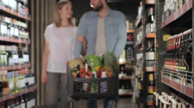 Çekici bir beyaz çift süpermarkette geziniyor, elleri alışveriş arabalarında taze ürünlere sürtünüyor. Bulanık arka plan renkli ürünlerle dolu canlı koridorları keşfederken neşeli bağlantılarını vurguluyor.