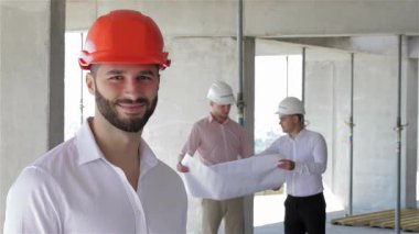 Gıcır gıcır beyaz gömlekli, parlak turuncu başlıklı, kendine güvenen genç, sakallı bir inşaat mühendisi. Arka planda, beyaz şapkalı iki inşaatçı odaklanmış bir tartışmaya girişiyor, planları gözden geçiriyorlar.