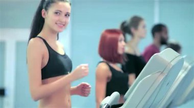 Zinde ve narin bir kız koşu bandında enerjik bir şekilde koşar, kendine güvenen bir gülümseme ve kameraya başparmak işareti yapar. Şık spor kıyafetleri giyip, spor salonu ortamında canlı bir motivasyon ve zindeliğin somutlaştırılmış hali.