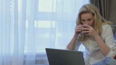 İnce, güzel bir kadın rahat bir koltukta oturur ve dizüstü bilgisayarına odaklanarak çayını yudumlar. Yumuşak doğal ışığın katıksız perdelerden süzülmesiyle, gevşemiş tavrı neşeli ve davetkar bir atmosfer yayıyor.