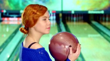 Neşeli, kızıl saçlı, neşeli, beyaz bir kızın yakın çekimi. Atışını yapmaya hazırlanırken elinde bir bowling topu tutuyor. Parlak gülümsemesi heyecan saçıyor. Oyunun heyecanını canlı bir bowling salonunda somutlaştırıyor.