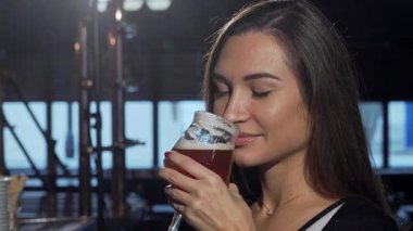 Uzun, dalgalı saçları olan çekici bir kadın ferahlatıcı bir zanaat birasından zevk alıyor. Gülümsemesi, düşünceli bir şekilde uzaktan bakarken neşe saçıyor. Barın sıcak atmosferi, kırsal ahşap barı ve yumuşak ışıklığıyla rahatlatıcı atmosferi arttırır.