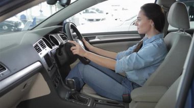 Şık, genç, beyaz bir kadın bayilikte modern bir arabanın sürücü koltuğunda rahatça oturur ve emniyet kemerini gülümseyerek bağlar. Davetkar iç mekan parlak bir gösterge paneli ve lüks deri koltuklar içeriyor.