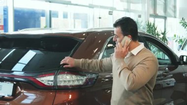 Koyu saçlı, kendine güvenen bir adam parlak bir araba kuaföründe şık, modern bir aracın yanında duruyor ve cep telefonuyla seçenekleri tartışıyor. Şık bir bej kazak ve siyah pantolon giyerek profesyonellik ve coşku saçar.