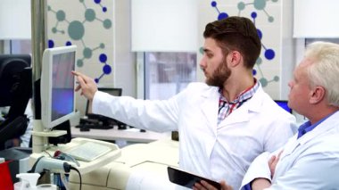 Şık bir sakalı olan odaklanmış genç bir beyaz adam, son teknoloji bir laboratuvardaki kaliteli bir bilgisayar ve tabletteki verileri karşılaştırıyor. Yanında, gri saçlı kıdemli bir laboratuar çalışanı düşünceli bir şekilde başını sallıyor ve dinamik bir işbirliği sahnesi yaratıyor.