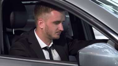 Genç bir işadamı şık, modern bir arabada kameraya bakarak rahatça otururken kendine güvenir. Bu görüntü lüks ve hırsın özünü yakalar, yeni otomotiv olanaklarını keşfetmenin heyecanını gözler önüne serer.