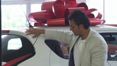 Şık, açık renk bir takım elbise giymiş neşeli, olgun bir adam yepyeni arabasının yanında şenlikli kırmızı bir fiyonkla poz verirken mutlulukla parıldıyor. Bu canlı sahne, tüketiciliğin heyecanını ve yeni sahipliğin sevincini yansıtıyor.