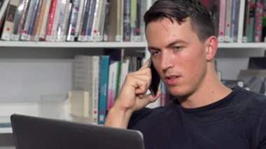 Çok odaklanmış yakışıklı bir adam, canlı bir üniversite kütüphanesinde dizüstü bilgisayarı üzerinde çalışırken bir telefon görüşmesi yapıyor. Kitap raflarıyla çevrelenmiş, modern eğitimin özünü somutlaştırıyor, teknolojiyi akademik arayışla harmanlıyor.