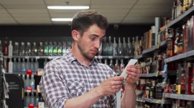 Şok olmuş bir ifadeye sahip genç bir adam, uzun alışveriş faturasını bir süpermarketin canlı koridorlarında inceliyor. Satın aldıklarının yüksek maliyetlerinden etkilenerek, canlı bir perakende piyasasında tüketiciliğin duygusal ağırlığını somutlaştırıyor.