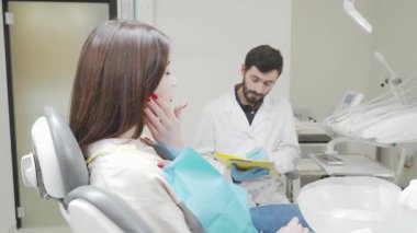 Acılı bir ifadeye sahip genç bir kadın, diş ağrısı çekerken yanağını nazikçe tutarak modern bir diş sandalyesine oturur. Beyaz önlük giymiş özenli dişçi, belgeleri doldurmaya odaklanmış durumda.