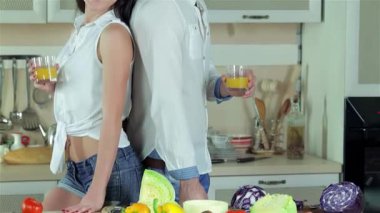 Neşeli, yeni evli bir çift parlak bir mutfakta sırt sırta dururlar, her biri ferahlatıcı bir bardak portakal suyu içerler. Canlı, taze kesilmiş sebzelerle çevrelenmiş olarak, birlikte yemek pişirme sevincini somutlaştırarak, canlı bir atmosfer yaratıyorlar.