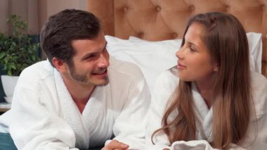 Neşeli bir çift, yumuşak bornozlara sarılmış lüks bir otel yatağında birlikte yatıyor, kahkahalar ve tatlı sohbetler ediyorlar. Sıcak gülümsemeleri ve şefkatli bakışları balaylarında romantik bir kaçamak için mükemmel bir ortam yaratıyor.