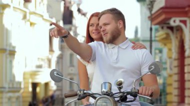 Neşeli genç bir çift birlikte scooter 'a binerken, erkek uzaktan bir şeyi işaret ederken kadın sıcak bir şekilde gülümsüyor ve onu kucaklıyor. Bu canlı şehir manzarası macera ve aşkın özünü yakalıyor, tasasız hallerini sergiliyor.