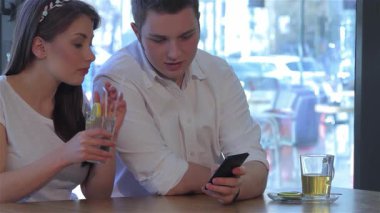 Saçında karışık bir halkayla çekici bir kadın akıllı telefonuna takılmış yakışıklı beyaz bir adamın yanında pipetle ferahlatıcı bir kokteyl yudumluyor. Konforlu kafe atmosferi pencereden süzülen yumuşak doğal ışık ile güçlendirilir.